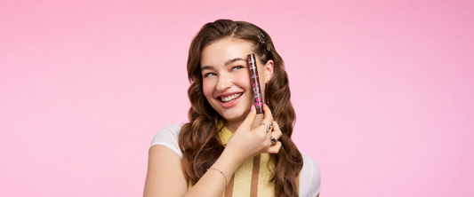 A woman holding a lash without limits tubing mascara in shade brown behind a pink background. 