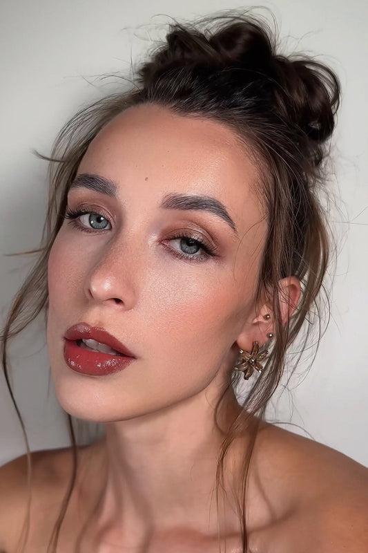 Woman with dark hair in a messy bun wearing gold earrings, posing with soft glam makeup.