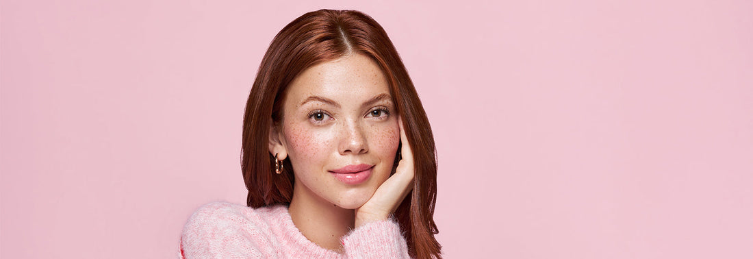 A women smiling with her hand on her chin behind a light pink background.