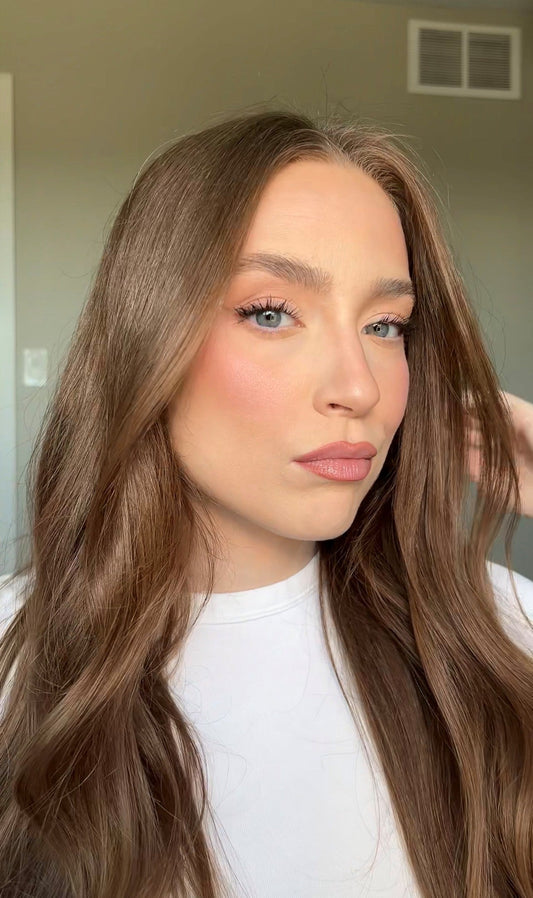 Woman with long wavy brown hair wearing a white top, posing with soft glam makeup.