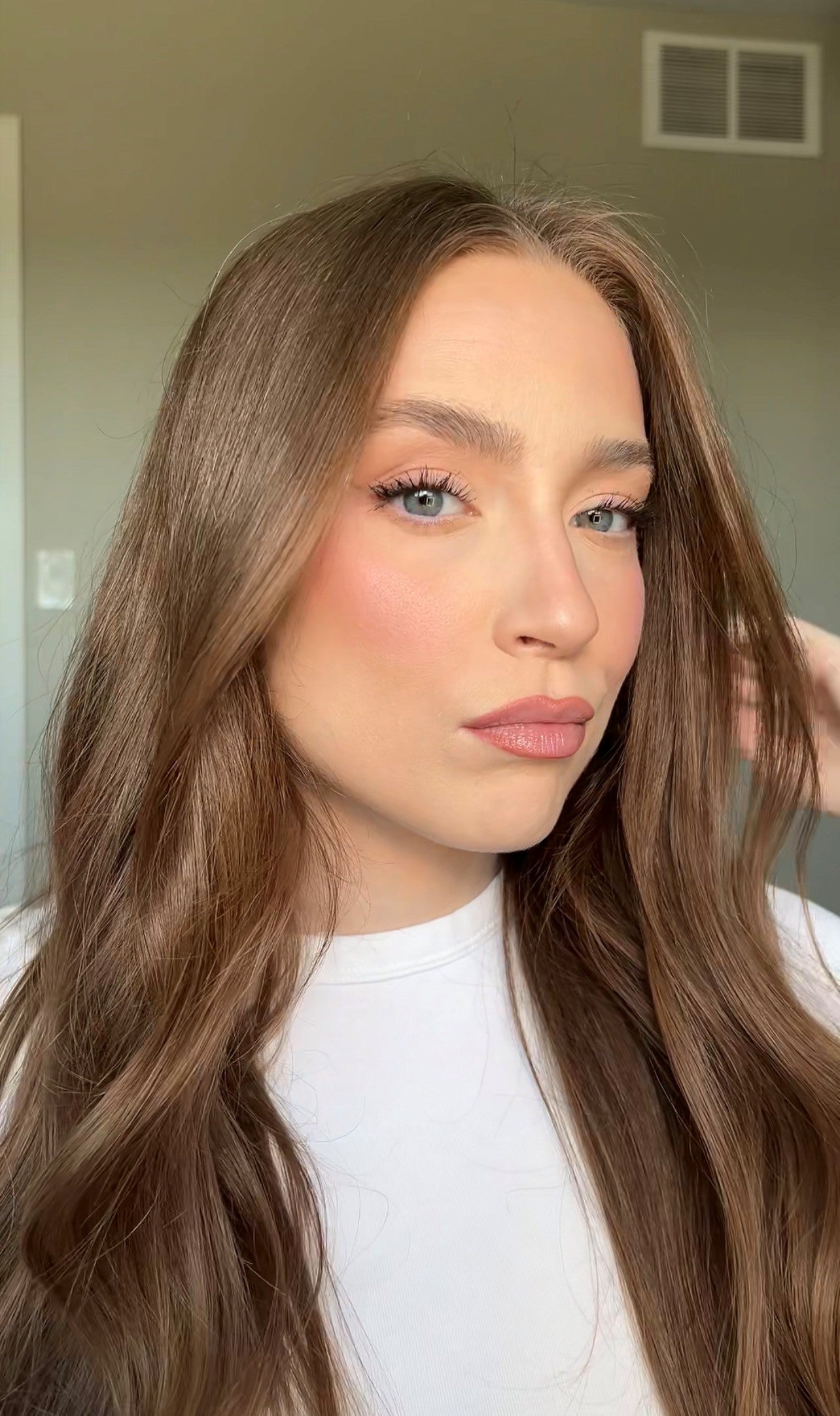 Woman with long wavy brown hair wearing a white top, posing with soft glam makeup.
