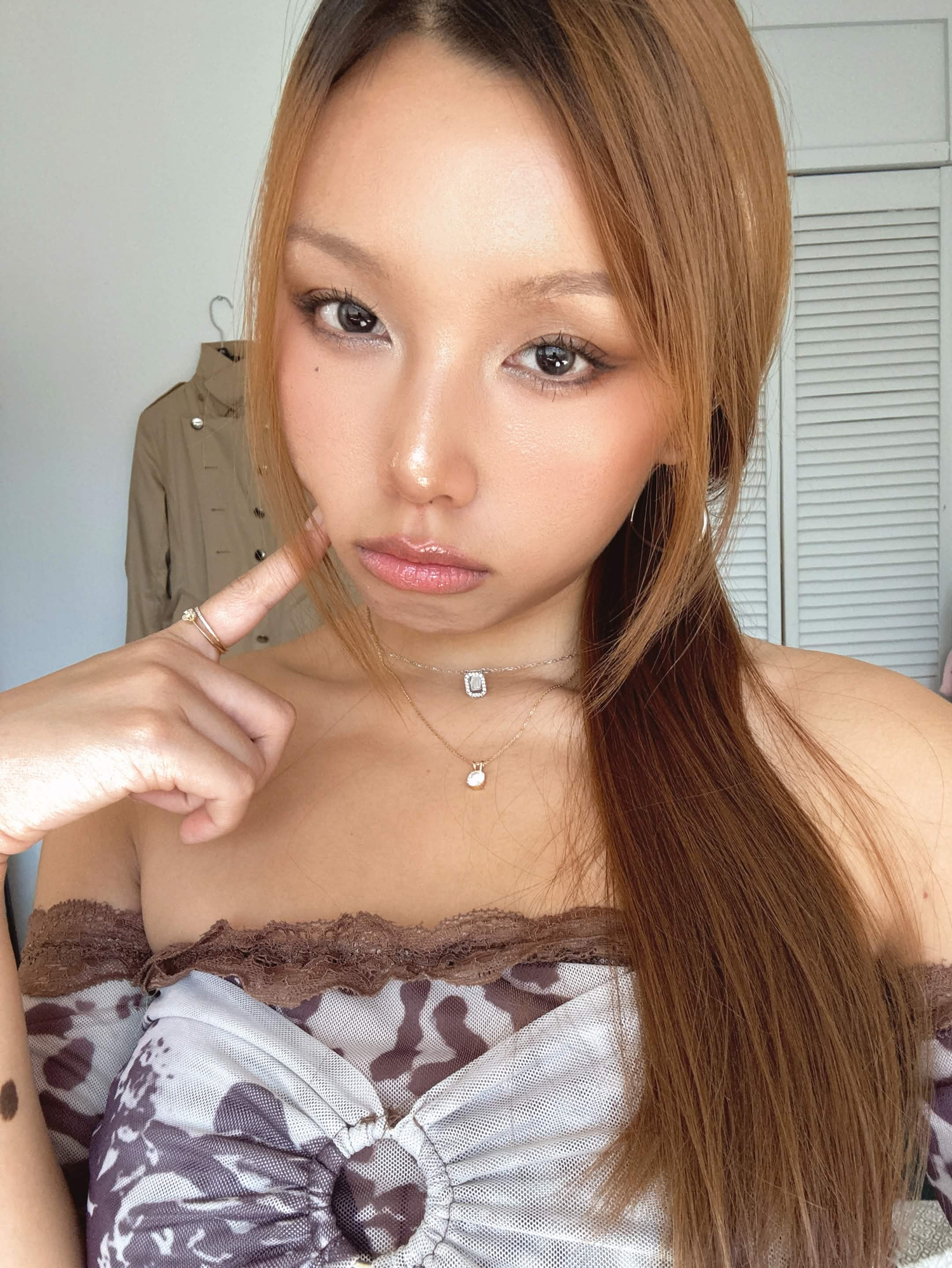 Young woman with long brown hair wearing an off-the-shoulder patterned top, posing with her finger on her cheek.