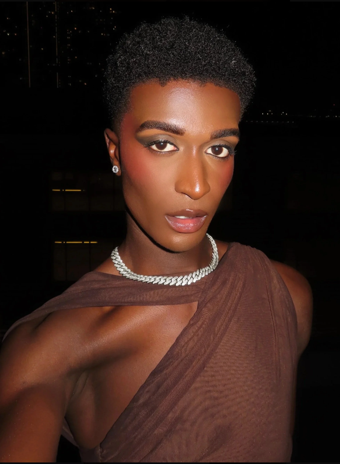 Person with short natural hair wearing a sheer brown one-shoulder top and pearl necklace, posing against a dark background.